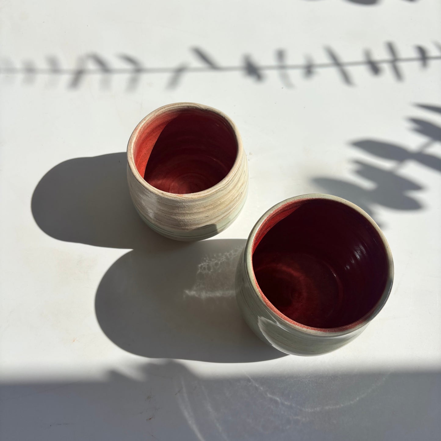 Two ceramic cups with red interior on a white surface with leaf shadows