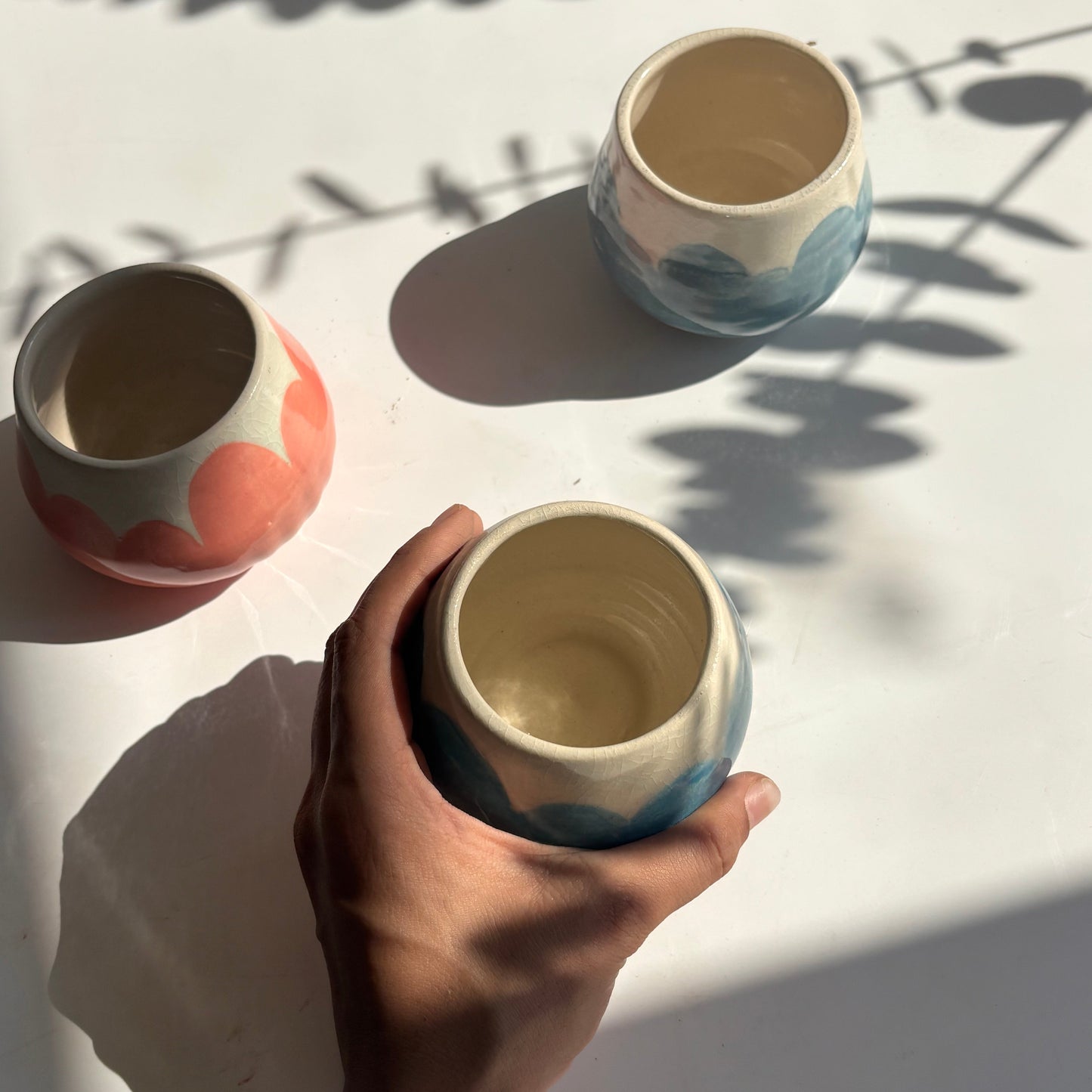 Hand holding a ceramic cup with another cup on a white surface, casting shadows.