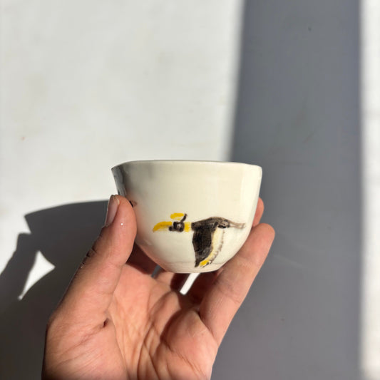 Hand holding a small white cup with black and yellow design against a light gray background