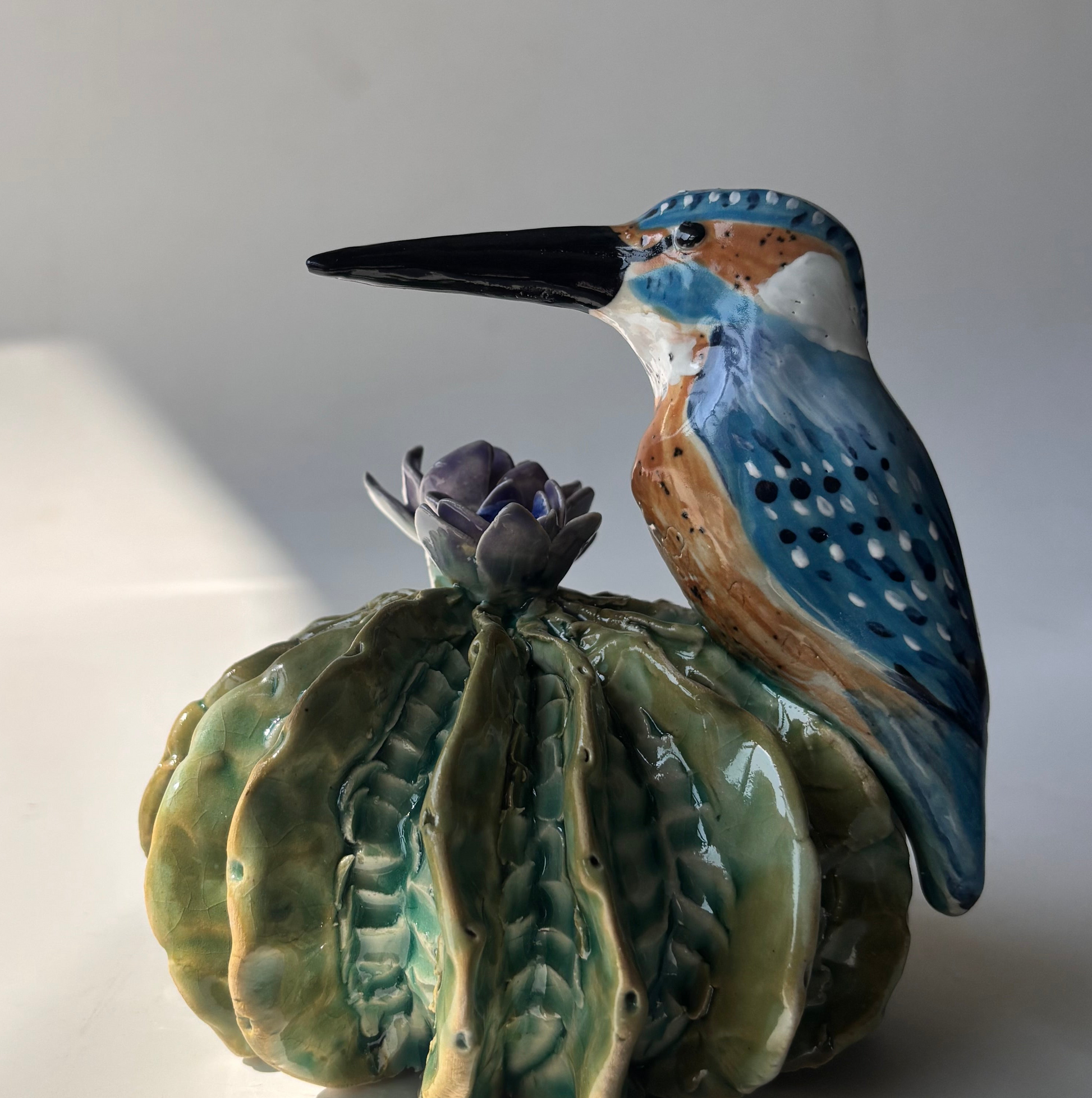 Ceramic kingfisher bird perched on a cactus with a purple flower against a white background