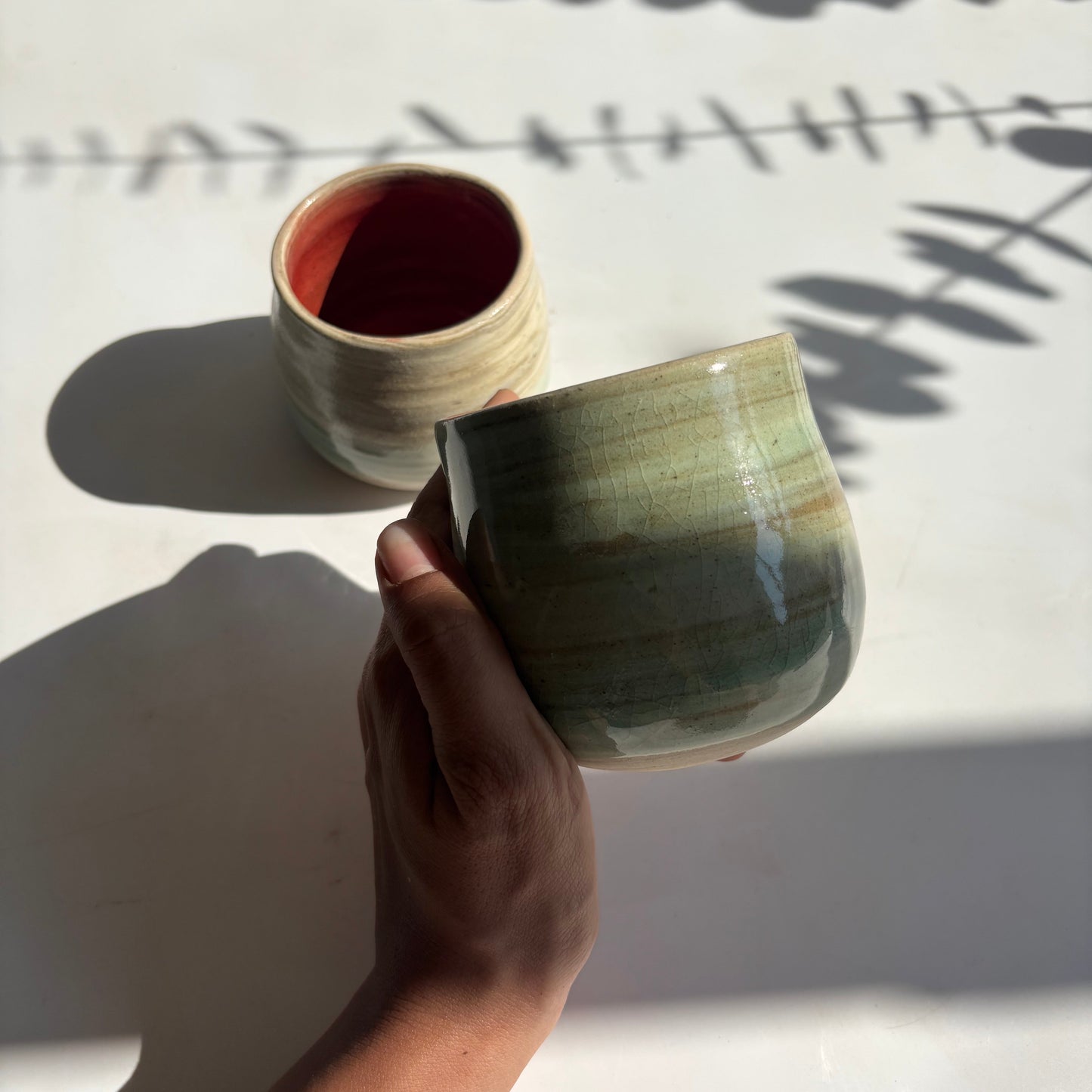 Hand holding a green ceramic cup with another cup in the background on a light surface.